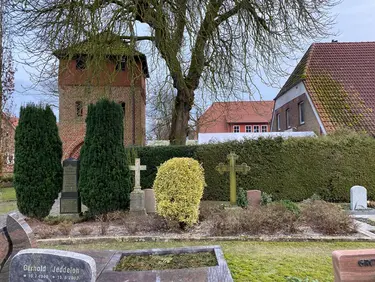 Im Vordergrund des Bildes sind bepflanzte und gepflegte Gräber mit Grabeinfassungen und Grabsteinen zu sehen. Durch das lichte Gebüsch hindurch sieht man den Glockenturm, der das Wahrzeichen Wardenburgs ist.  